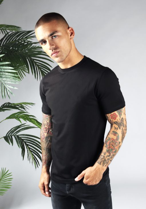 Linker zijaanzicht heren T-shirt in zwarte kleur en een straight fit pasvorm. Het model draagt een donkere jeans en heeft zijn linkerarm in zijn linker broekzak.