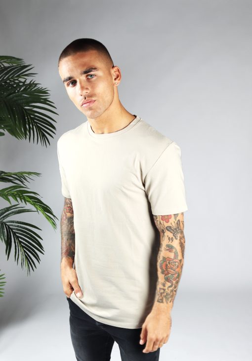 Linker vooraanzicht heren T-shirt in sand kleur en een long fit pasvorm. Het model draagt een donkere jeans en heeft zijn rechterhand in zijn rechter broekzak.