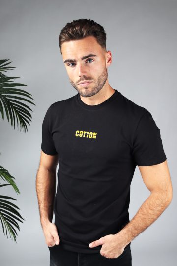 Vooraanzicht heren T-shirt in zwarte kleur en een straight fit pasvorm. Het T-shirt is voorzien van de tekst 'COTTON' in het geel. Het model heeft beiden handen in zijn broekzakken.