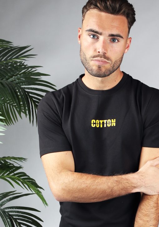Vooraanzicht heren T-shirt in zwarte kleur en straight fit pasvorm. Het T-shirt is voorzien van de tekst 'COTTON' in het geel. Het model heeft zijn linkerarm vast met zijn rechterarm.