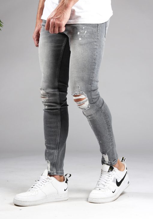 Linker zijkant lichtgrijze denim heren skinny jeans met damaged look, scheuren en verfspatten. Gecombineerd met wit T-shirt en witte sneakers.