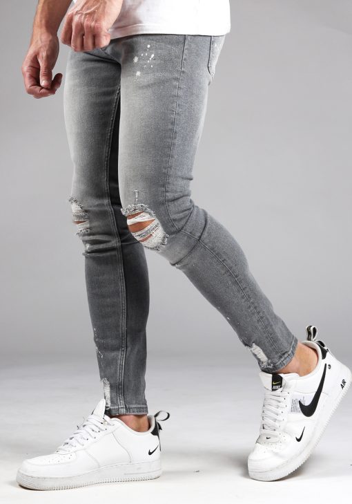 Linker zijkant lichtgrijze denim heren skinny jeans met damaged look, scheuren en verfspatten. Gecombineerd met wit T-shirt en witte sneakers.