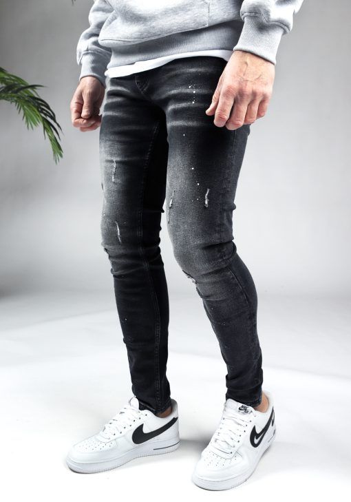 Linker zijaanzicht zwarte heren skinny jeans met verfspetters en kleine scheuren, gemaakt van stretch stof. Voorzien van vier zakken, knoopsluiting en riemlussen. Gecombineerd met grijze hoodie en witte sneakers.