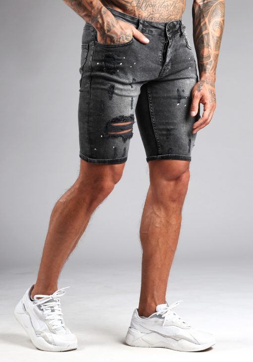 Rechter vooraanzicht van model gekleed in grijze jean shorts met damaged details en verfspetters. Het model heeft een hand in zijn broekzak en draagt witte sneakers.
