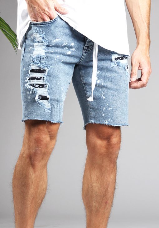 Rechter vooraanzicht van model gekleed in blauwe jean shorts in combinatie met een wit shirt. De shorts hebben witte verfspetters en gaten waaronder een zwart witte rodeo print te zien is. Ook loopt er een wit touwtje door de riemgaten van de jeans. Het model heeft een hand in zijn zak.