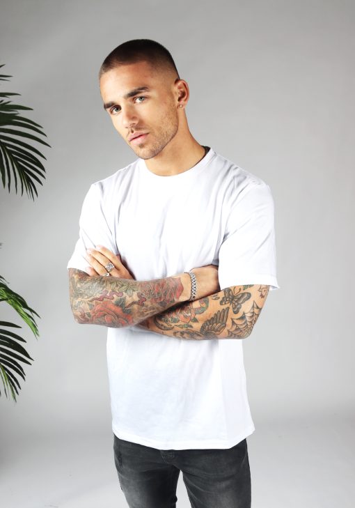 Vooraanzicht van heren T-shirt in witte kleur, met ronde hals en oversized pasvorm. Het model draagt een donkere jeans en heeft zijn armen over elkaar.