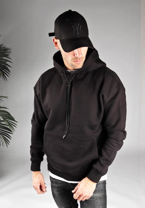 Vooraanzicht van model gekleed in een zwarte hoodie met lange hoodie strings in combinatie met een zwarte broek. Het model draagt een zwarte pet, heeft zijn armen losjes naast zich, en kijkt schuin naar de grond.