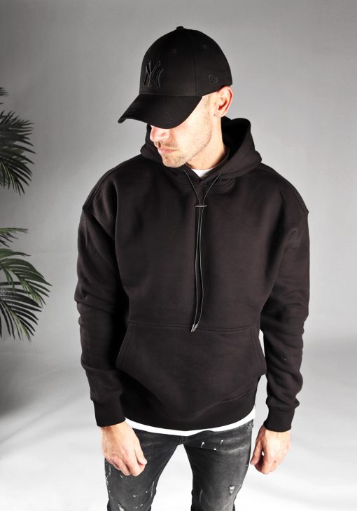 Vooraanzicht van model gekleed in een zwarte hoodie met lange hoodie strings in combinatie met een zwarte broek. Het model draagt een zwarte pet, heeft zijn armen losjes naast zich, en kijkt langs zijn rechter schouder naar de grond.