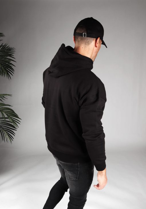 Schuin achteraanzicht van model gekleed in een zwarte hoodie in combinatie met een zwarte broek. Het model draagt een zwarte pet, heeft zijn armen losjes naast zich, en kijkt naar de grond.