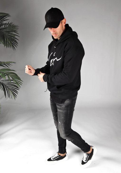 Zijaanzicht van model gekleed in een zwarte hoodie met lange hoodie strings en het grote witte signature cotton logo op de borst gecombineerd met een zwarte broek. Het model draagt een zwarte pet, heeft zijn handen voor zich, en kijkt schuin naar de grond.