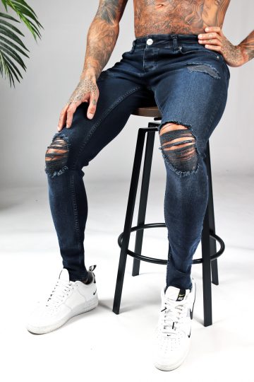 Man met donkerblauwe denim heren skinny jeans met damaged look, scheuren en verfspatten zit op een kruk.