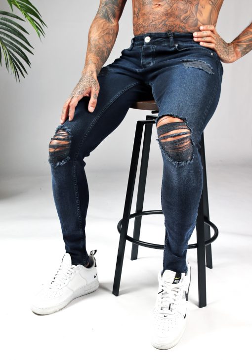 Man met donkerblauwe denim heren skinny jeans met damaged look, scheuren en verfspatten zit op een kruk.
