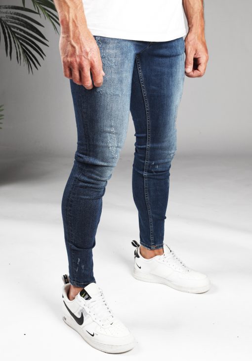 Rechter zijkant denim heren skinny jeans met damaged look, gemaakt van stretch stof. Voorzien van vier zakken, knoopsluiting en riemlussen. Gecombineerd met wit T-shirt en witte sneakers.