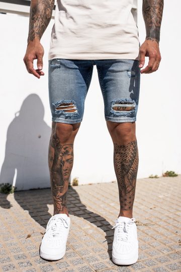 Vooraanzicht onderlichaam van model gekleed in blauwe jean shorts met damaged details in combinatie met witte sneakers en een beige top. Het model staat buiten voor een witte muur en heeft zijn armen losjes langs zich.