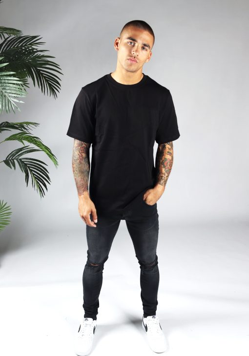 Vooraanzicht van heren T-shirt in zwarte kleur, met ronde hals en een dropshoulder fit pasvorm. Het model heeft zijn linkerhand in zijn linkerbroekzak, draagt een donkere jeans en witte sneakers.