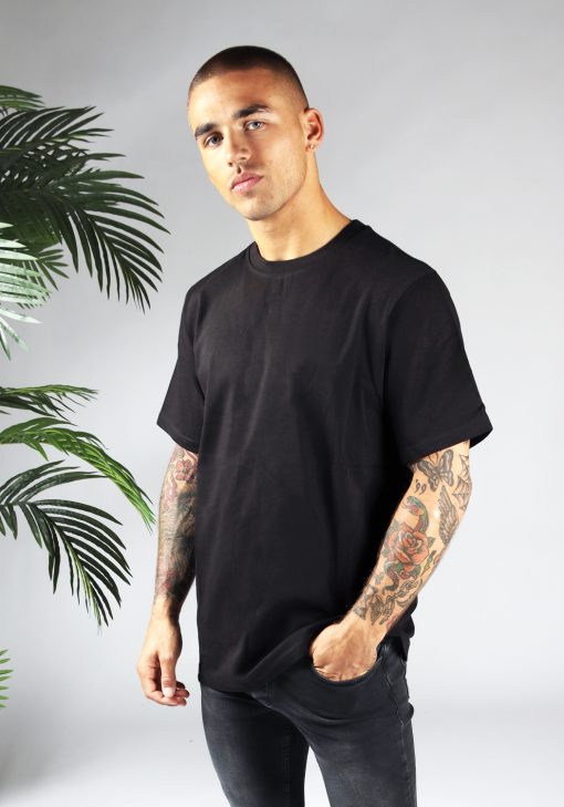 Linker zijaanzicht van heren T-shirt in zwarte kleur, met ronde hals en een dropshoulder fit pasvorm. Het model heeft zijn linkerhand in zijn linkerbroekzak.