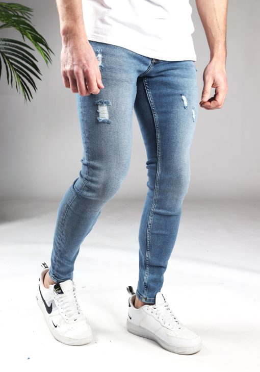 Rechter zijaanzicht blauwe heren jeans met skinny pasvorm en damaged look. Voorzien van twee broekzakken, twee kontzakken, knoopsluiting en riemlussen. Gecombineerd met wit T-shirt en witte sneakers.