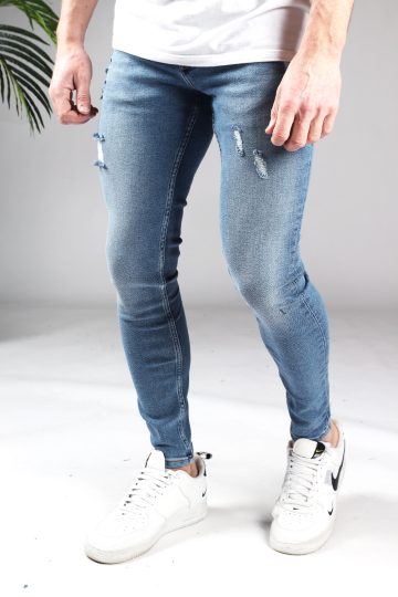 Vooraanzicht blauwe heren jeans met skinny pasvorm en damaged look. Voorzien van twee broekzakken, twee kontzakken, knoopsluiting en riemlussen. Gecombineerd met wit T-shirt en witte sneakers.