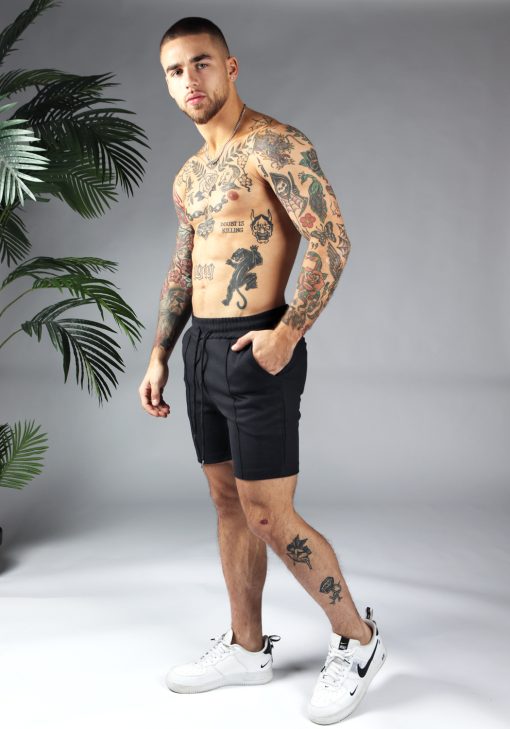 Linker zijaanzicht comfortabele heren chino shorts in zwarte kleur, met skinny fit en gemaakt van luxe stretch stof. Voorzien van twee broekzakken en rekbare taille met trekkoord. Gecombineerd met witte sneakers.