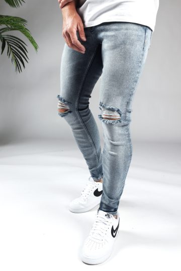 Linker zijkant lichte denim heren skinny jeans met gescheurde knieën, gemaakt van stretch stof. Voorzien van twee zakken aan de voorzijde, twee kontzakken, knoopsluiting en riemlussen. Gecombineerd met wit T-shirt en witte sneakers.
