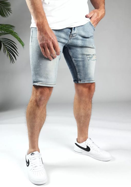 Schuin rechter vooraanzicht van model gekleed in lichtblauwe trench jean shorts. De shorts hebben damaged details en een skinny fit. Het model heeft een hand in de broekzak.