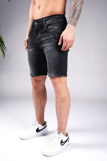 Linker zijaanzicht van model gekleed in zwarte skinny-relaxed jean shorts met verf spetter details. Het model heeft zijn handen naast zich en draagt witte sneakers.