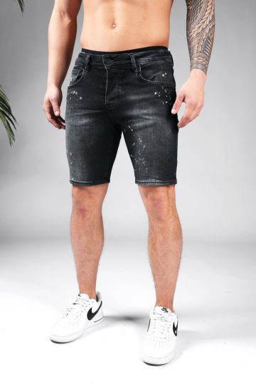Vooraanzicht van model gekleed in zwarte skinny-relaxed jean shorts met verf spetter details. Het model heeft zijn handen naast zich en draagt witte sneakers.