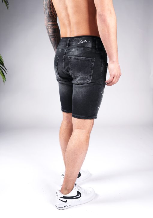 Schuin achteraanzicht van model gekleed in zwarte skinny-relaxed jean shorts met verf spetter details. Het model heeft zijn handen naast zich en draagt witte sneakers.
