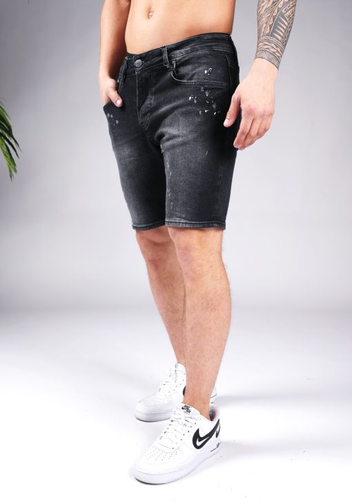 Linker zijaanzicht van model gekleed in zwarte skinny-relaxed jean shorts met verf spetter details. Het model heeft een hand in zijn zak en draagt witte sneakers.