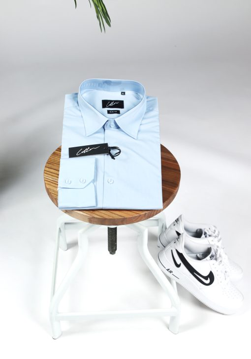 Opgevouwen heren overhemd in een blauwe kleur, met zwarte knoopjes en een kledinglabel. Het overhemd ligt op een kruk en er liggen witte sneakers tegen de kruk aan.