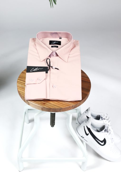 Opgevouwen heren overhemd in een roze kleur, met roze knoopjes en een kledinglabel. Het overhemd ligt op een kruk en er liggen witte sneakers tegen de kruk aan.
