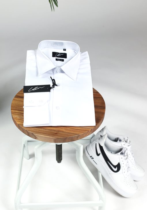 Opgevouwen heren overhemd in een witte kleur, met witte knoopjes en een kledinglabel. Het overhemd ligt op een kruk en er liggen witte sneakers tegen de kruk aan.