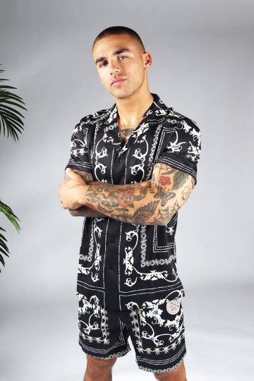Linker zijaanzicht van model gekleed in zwarte set met witte hawai print. De set bestaat uit shorts en een blouse met korte mouwen. Het model heeft zijn armen voor zich over elkaar.