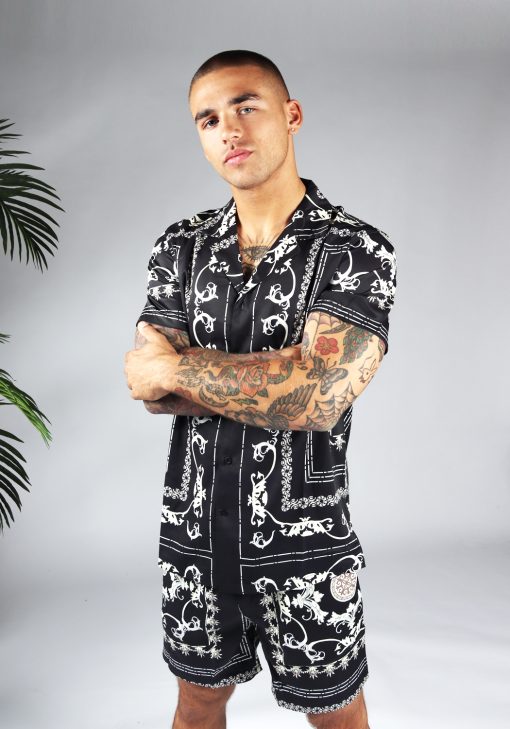 Linker zijaanzicht van model gekleed in zwarte set met witte hawai print. De set bestaat uit shorts en een blouse met korte mouwen. Het model heeft zijn armen voor zich over elkaar.