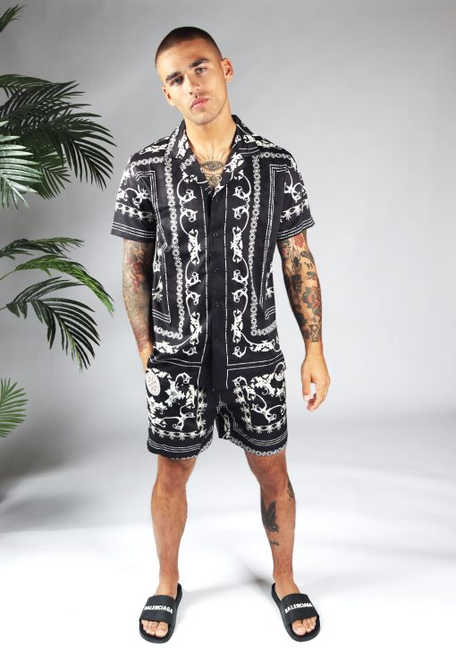Vooraanzicht van model gekleed in zwarte set met witte hawai print. De set bestaat uit shorts en een blouse met korte mouwen. Het model heeft een hand in zijn broekzak.
