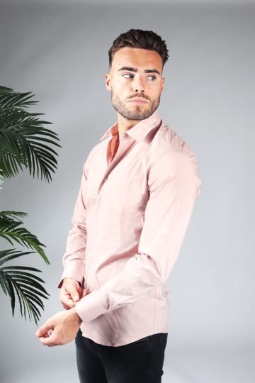 Linker zijaanzicht van heren overhemd in een roze kleur, met roze knoopjes en een slimfit pasvorm. Het model draagt een donkere jeans en heeft zijn linkermouw vast met zijn rechterhand.