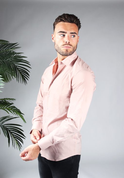 Linker zijaanzicht van heren overhemd in een roze kleur, met roze knoopjes en een slimfit pasvorm. Het model draagt een donkere jeans en heeft zijn linkermouw vast met zijn rechterhand.