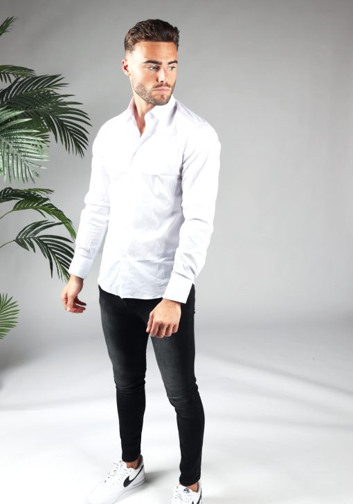 Vooraanzicht van heren overhemd in een witte kleur, met witte knoopjes en een slimfit pasvorm. Het model draagt een donkere jeans, witte sneaker en kijkt naar links.