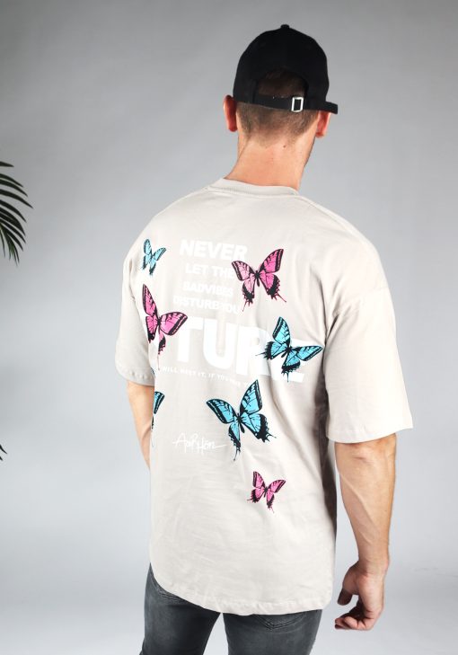 Schuin achteraanzicht van heren T-shirt in taupe kleur, met ronde hals en een oversized pasvorm. Het T-shirt is voorzien van vlinder print in roze en blauwe kleur en witte tekst. Het model draagt een zwarte pet en donkere jeans.
