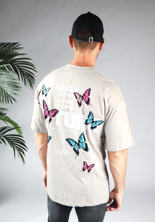 Achteraanzicht van heren T-shirt in taupe kleur, met ronde hals en een oversized pasvorm. Het T-shirt is voorzien van vlinder print in roze en blauwe kleur en witte tekst. Het model draagt een zwarte pet en donkere jeans.