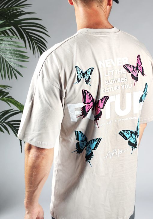 Close-up van heren T-shirt in taupe kleur, met ronde hals en een oversized pasvorm. Het T-shirt is voorzien van vlinder print in roze en blauwe kleur en witte tekst.
