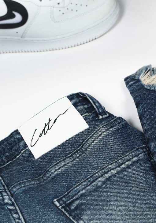 Close up Cotton-logo op de achterkant van de Cotton Side Zip jeans: blauwe heren skinny jeans met damaged uitstraling, scheuren in de broekspijpen en ritsen aan de zijkant van de benen.