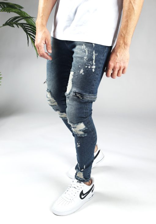 Linker zijkant blauwe heren skinny jeans met damaged uitstraling, scheuren in de broekspijpen en ritsen aan de zijkant van de benen. Voorzien van broekzakken, knoopsluiting en riemlussen.