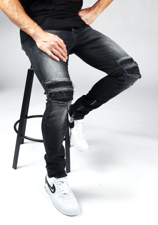 Man zit op kruk en draagt een zwarte heren jeans slim fit model met scheuren bij de knieën en een rits aan de binnenkant van de enkel.