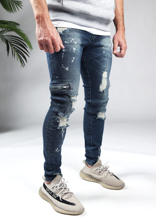 Rechter zijkant blauwe heren skinny jeans met damaged uitstraling, scheuren in de broekspijpen en ritsen aan de zijkant van de benen. Voorzien van broekzakken, knoopsluiting en riemlussen.