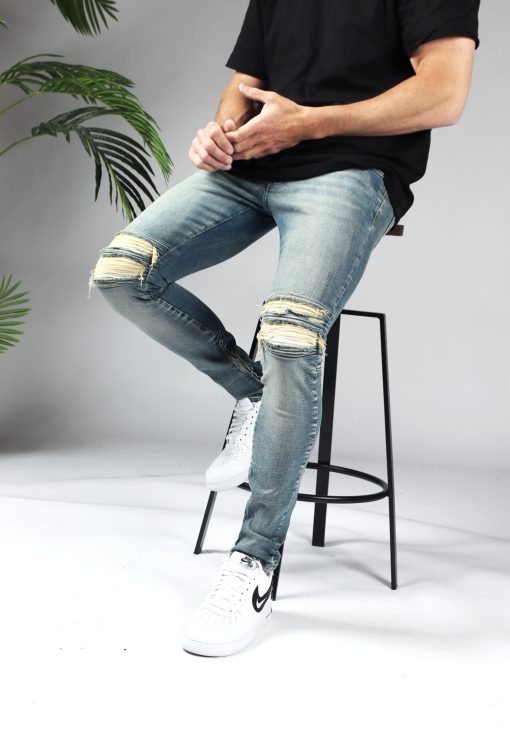 Linker zijaanzicht zittende man die een lichtblauwe heren jeans slim fit model met scheuren bij de knieën en een rits aan de binnenkant van de enkel draagt.