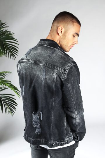 Schuin achteraanzicht van model gekleed in zwarte denim jacket met beschadigde details en zilveren drukknopen. Het model heeft zijn handen langs zich en kijkt schuin naar de grond.