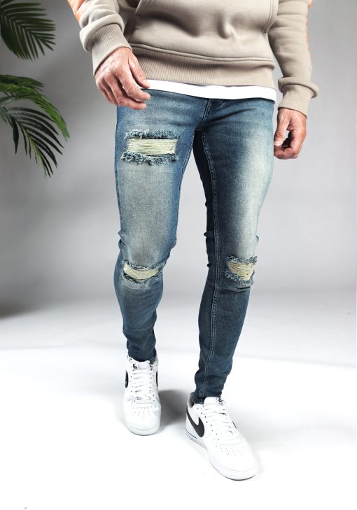 Voorkant blauwe heren skinny jeans met scheuren in de broekspijpen en gemaakt van stretch stof. Voorzien van vier zakken, knoopsluiting en riemlussen. Gecombineerd met crèmekleurige hoodie en witte sneakers.