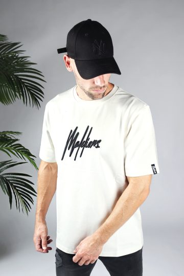 Vooraanzicht van model gekleed in gebroken wit shirt met drop shoulder fit en het zwarte Malelions logo op het midden van de borst. Aan de linkermouw zit een kleine zwarte tag met het witte logo van Malelions.