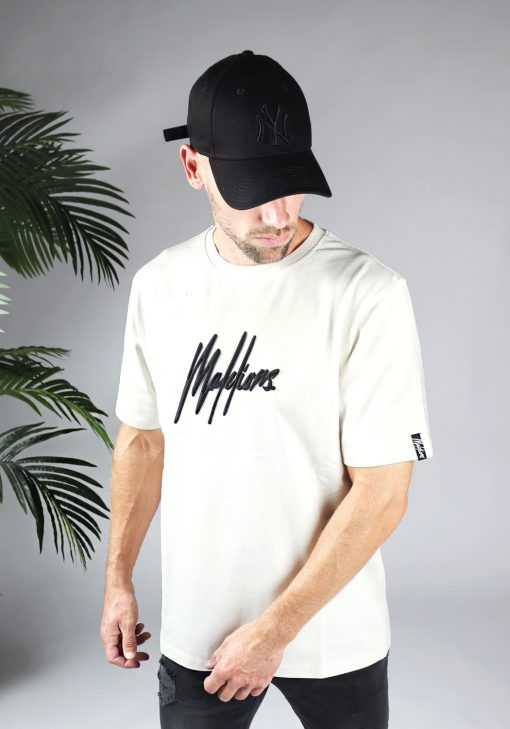 Vooraanzicht van model gekleed in gebroken wit shirt met drop shoulder fit en het zwarte Malelions logo op het midden van de borst. Aan de linkermouw zit een kleine zwarte tag met het witte logo van Malelions.